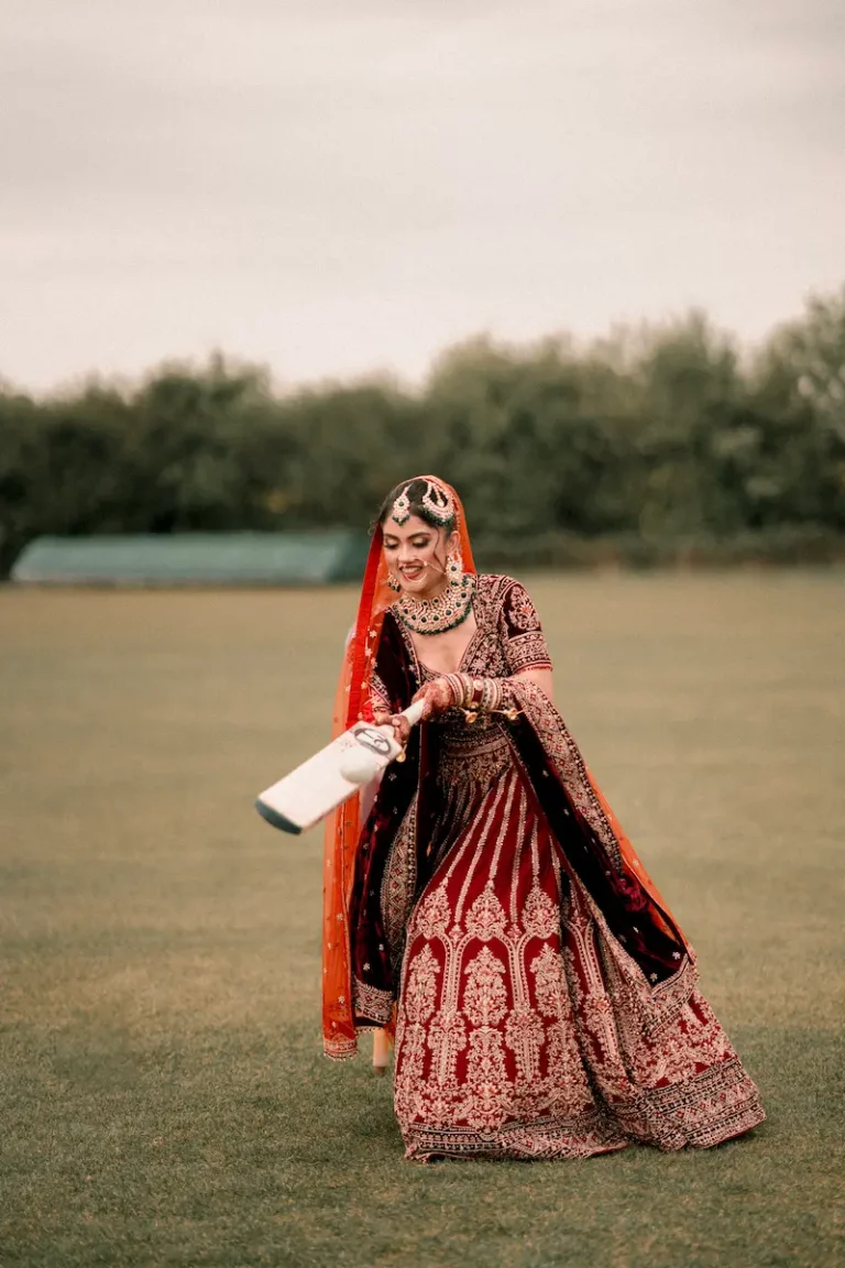 Bride in red bridal lehenga playfully swinging a cricket bat, candid wedding photography in London