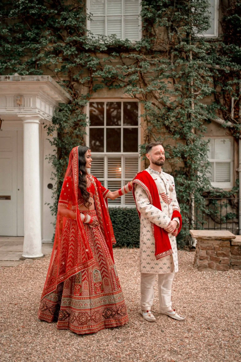Hindu bride in red and gold embroidered lehenga walking with groom in white sherwani, London wedding venue