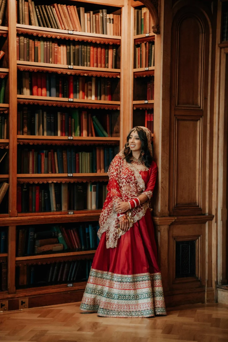 Hindu bride in red and gold embroidered lehenga standing beside library bookshelves