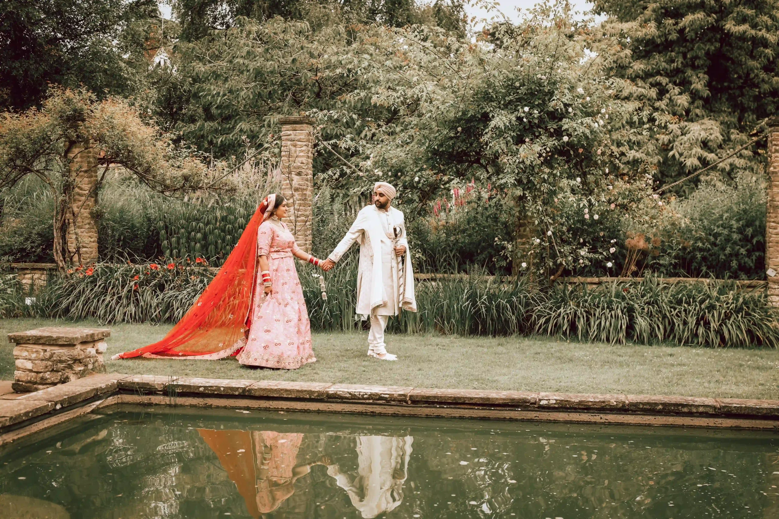Hindu bride in pink lehenga and groom in cream pagri walking beside garden pool