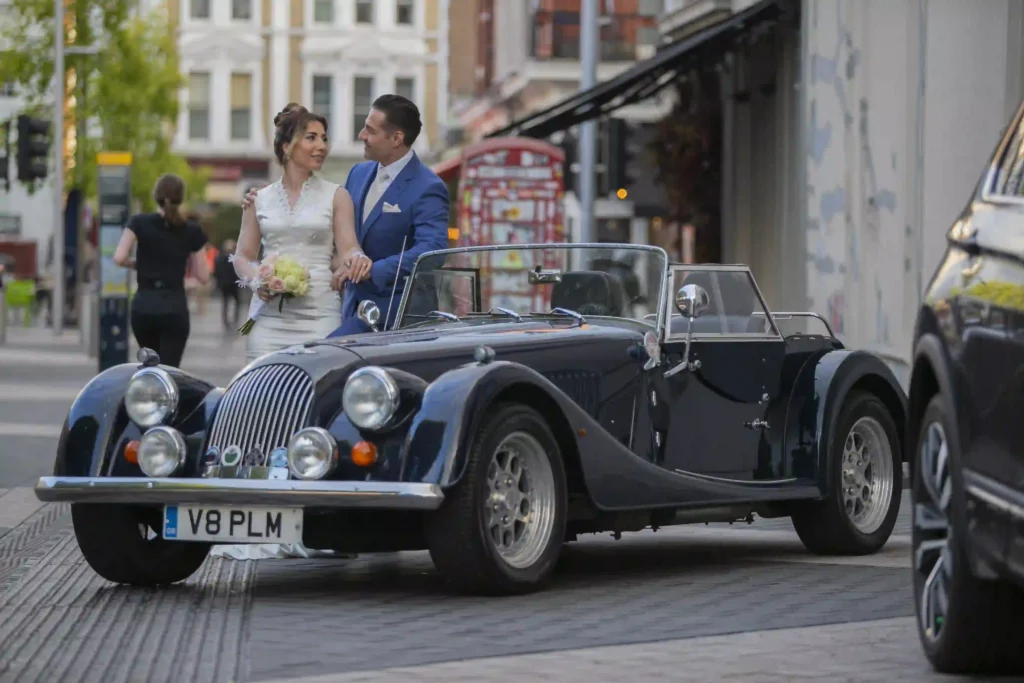 Bride and groom standing beside a vintage convertible car on a London street, candid wedding photography by Memory Filming