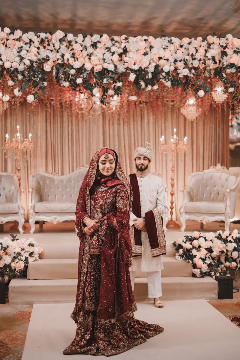 Pakistani bride in maroon dupatta and groom in a white pagri standing in front of a white rose floral stage backdrop for Baraat coverage