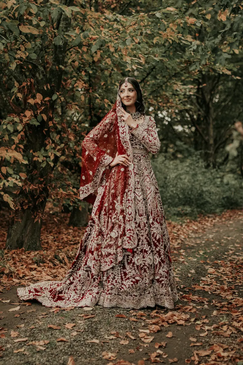 Full-length editorial portrait of a Pakistani bride in a red and gold suit standing along a tree-lined autumn path in a London park