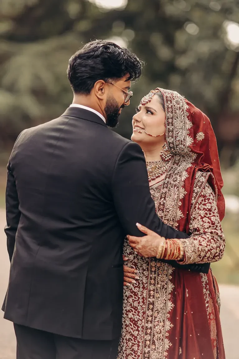 Close-up portrait of a groom in a black suit holding his bride in an ornate red and gold embroidered outfit outdoors