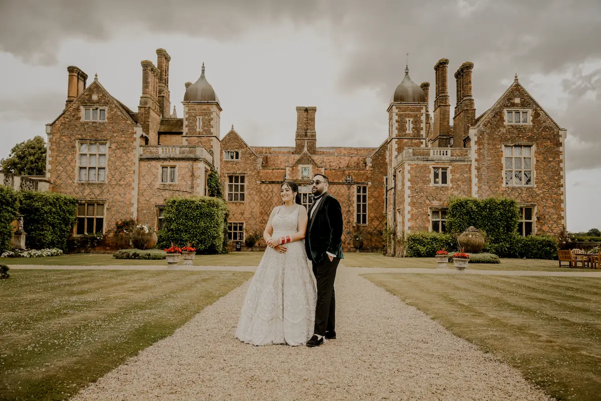 Groom in a velvet tuxedo jacket and bride in an embellished gown posing on a gravel pathway in front of a grand Tudor mansion capture by english wedding photographer london