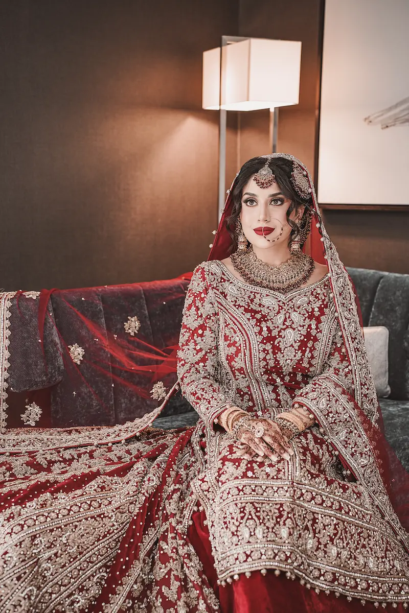 High-fashion Pakistani bridal photography showing a bride in a heavily embroidered red Barat outfit sitting gracefully indoors