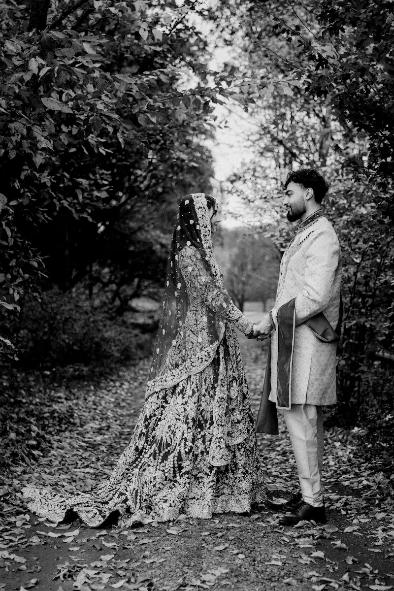 Black and white portrait of a bride and groom holding hands while looking at each other along a tree-lined autumn path in London