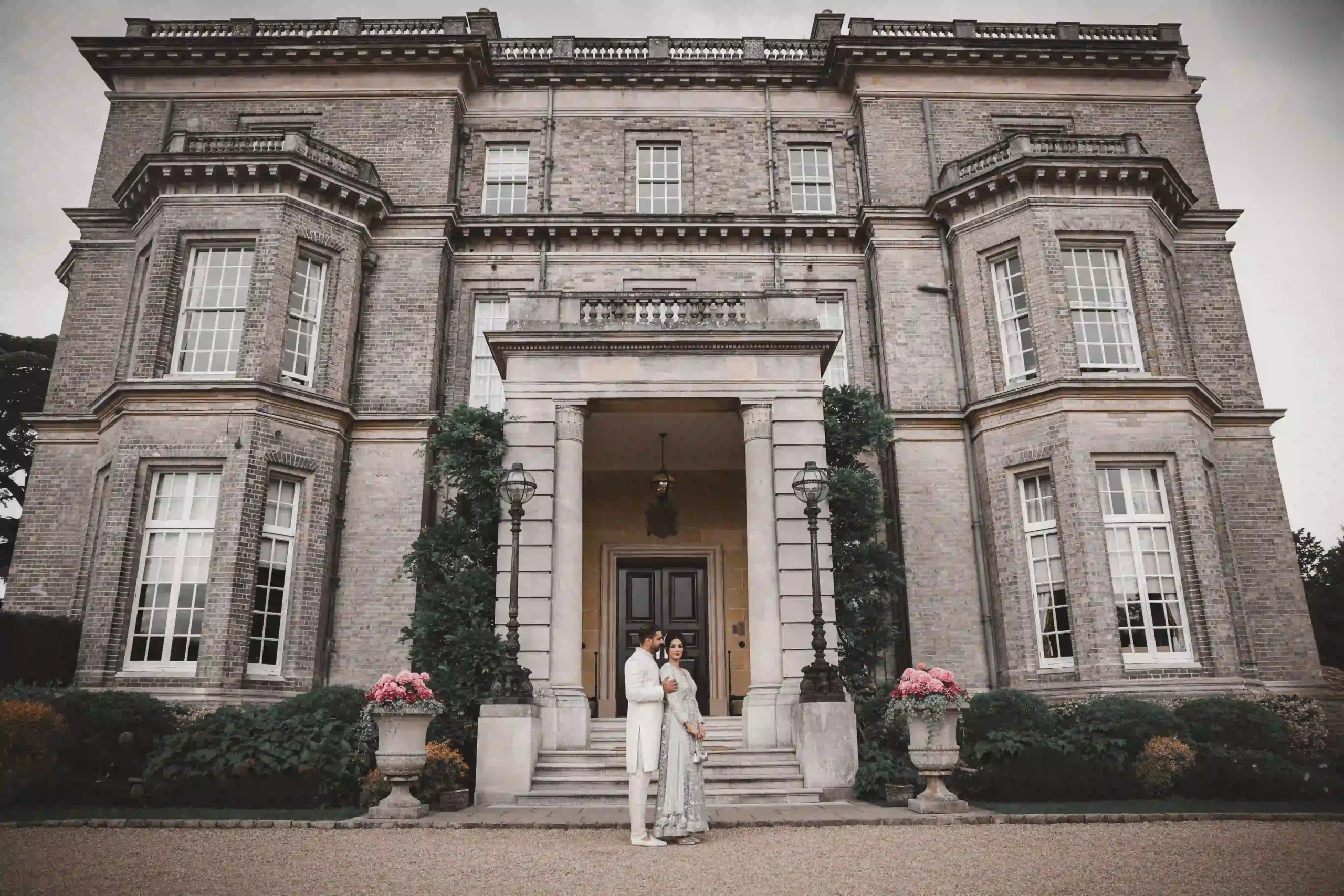 Couple in pastel wedding outfits standing at the stone entrance of a grand country estate documented by an Asian wedding photographer and videographer in the UK