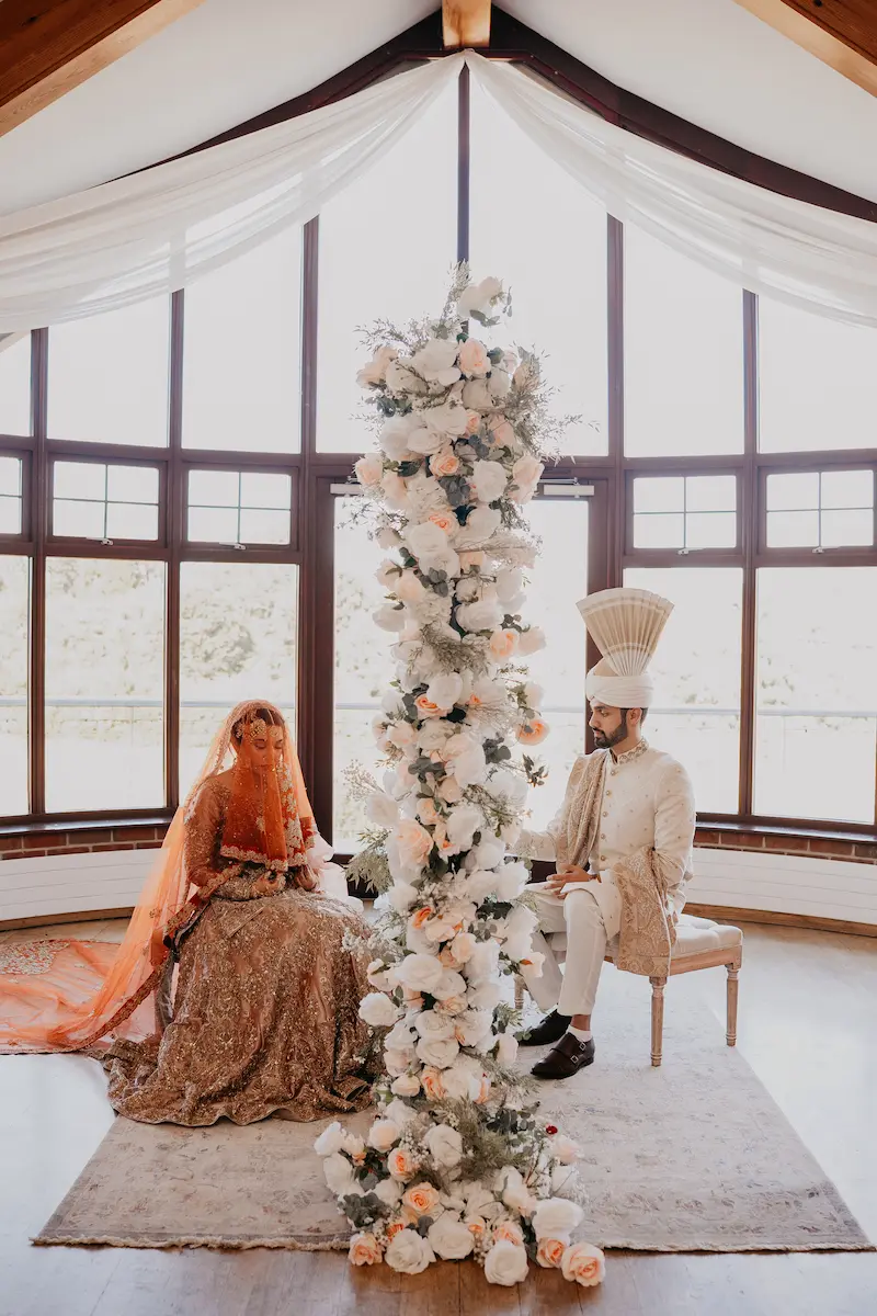 Pakistani wedding photographer capturing an intimate Nikkah photoshoot of a bride and groom seated on opposite sides of a white floral pillar