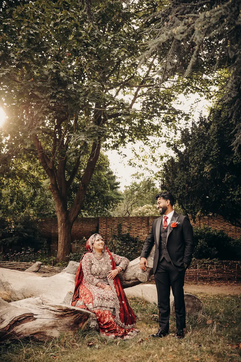 Pakistani wedding photo and video team capturing a couple portrait with the bride sitting on a fallen log and groom in a dark suit standing nearby
