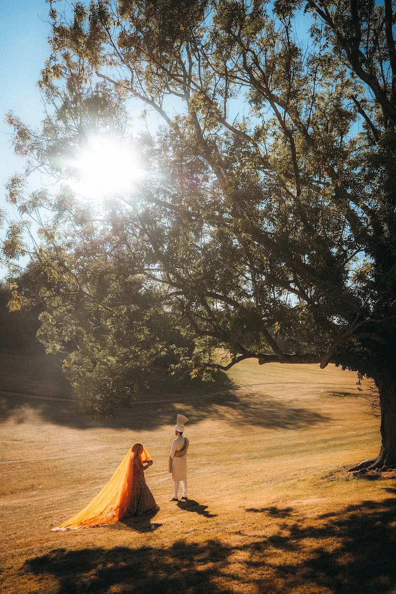 Cinematic Pakistani wedding photography of a bride in a long orange veil and groom in a white sherwani standing under a large oak tree during a golden hour photoshoot in London