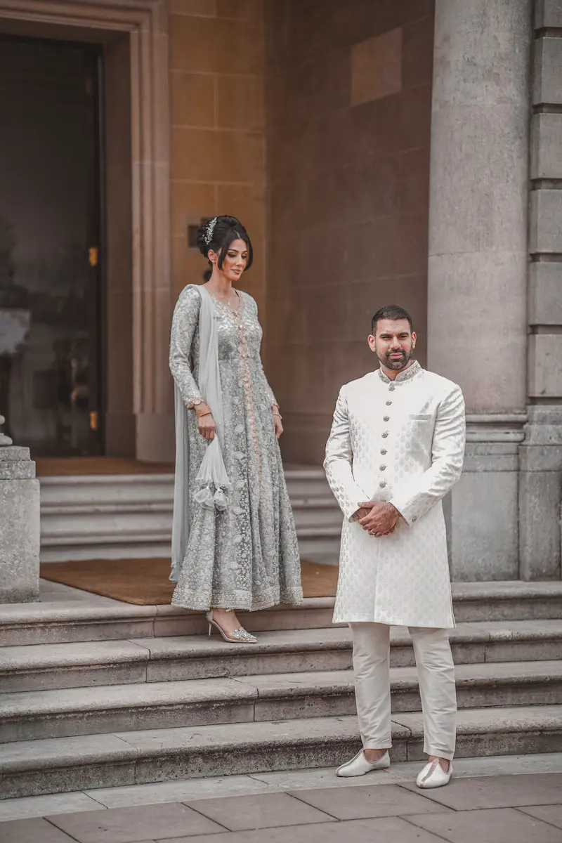 Bride in an embellished silver outfit standing on stone steps while groom in a cream sherwani stands below at a historic London estate