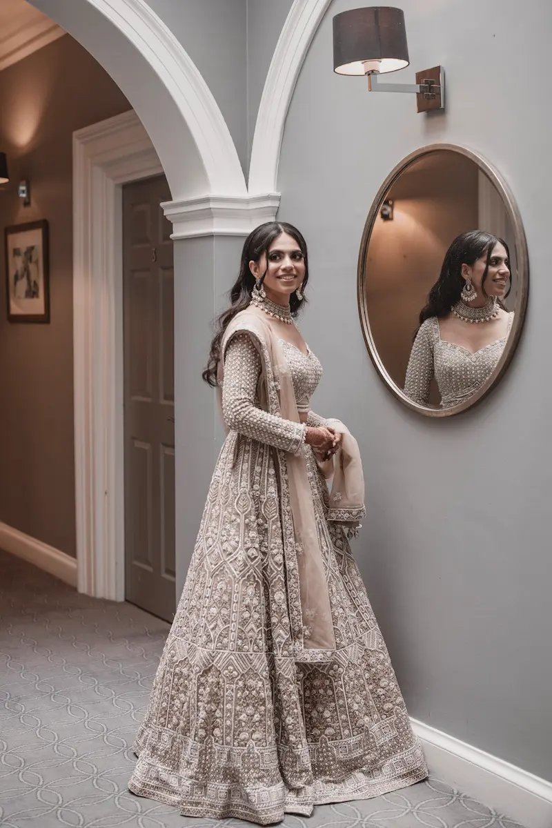 Bride in an intricately beaded silver-beige lehenga smiling next to a wall mirror in a luxury London venue hallway