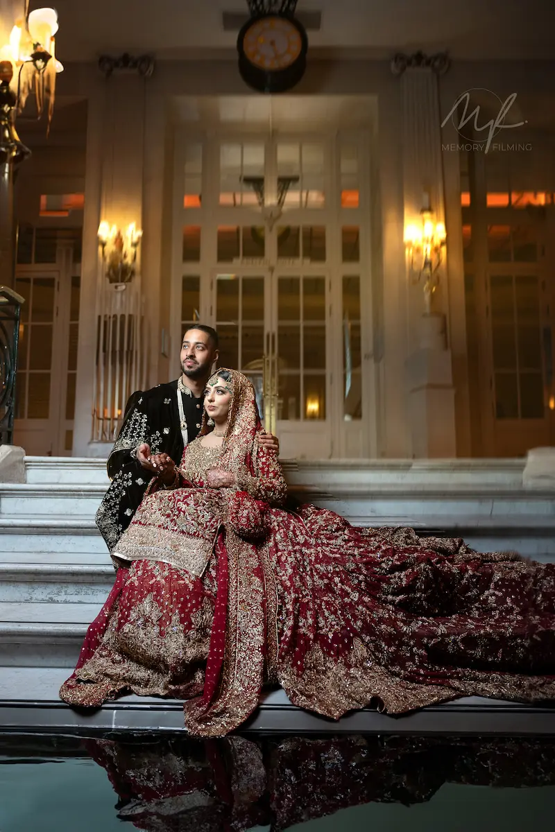 Couple in embroidered dark velvet and maroon regal attire sitting together on a grand white marble staircase at a London venue