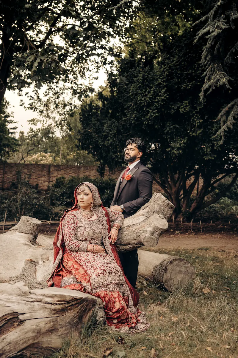 Bride in traditional heavily embroidered red attire sitting on a large fallen tree trunk while her groom in a suit stands beside her in a park