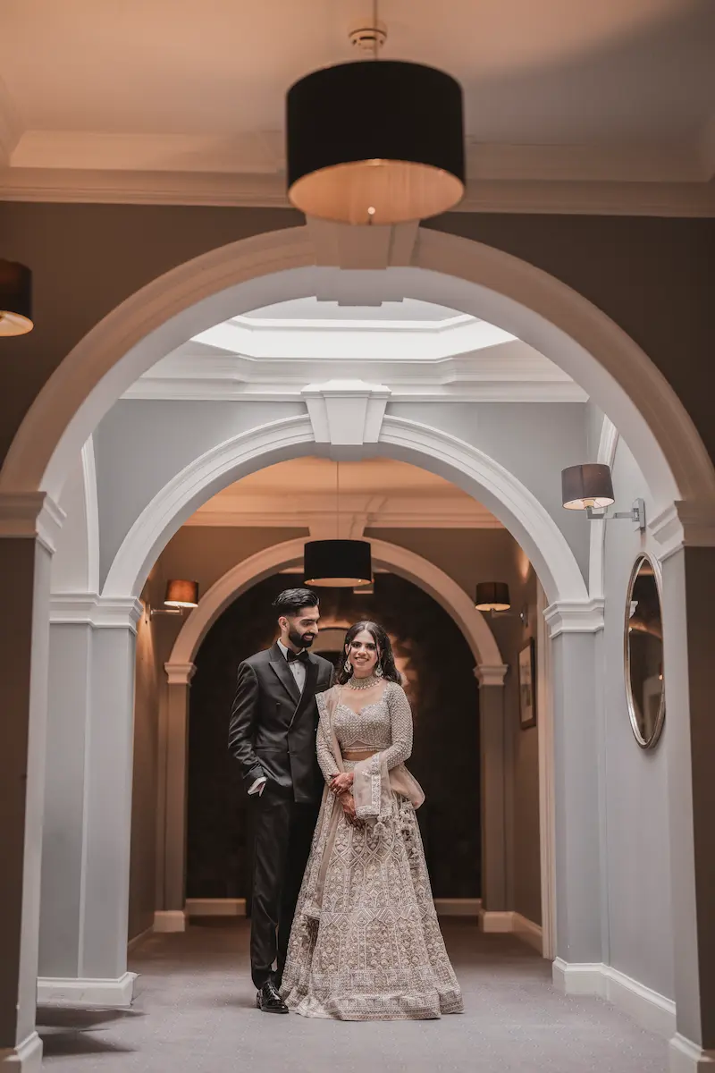 Pakistani couple in a black tuxedo and silver embellished lehenga posing in an arched hallway, shot by a professional Pakistani wedding photographer