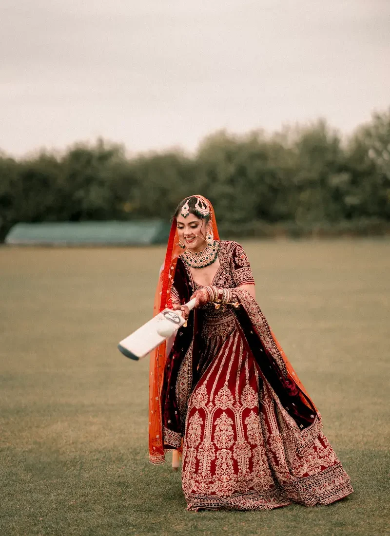 Bride in red bridal lehenga playfully swinging a cricket bat, candid wedding photography in London